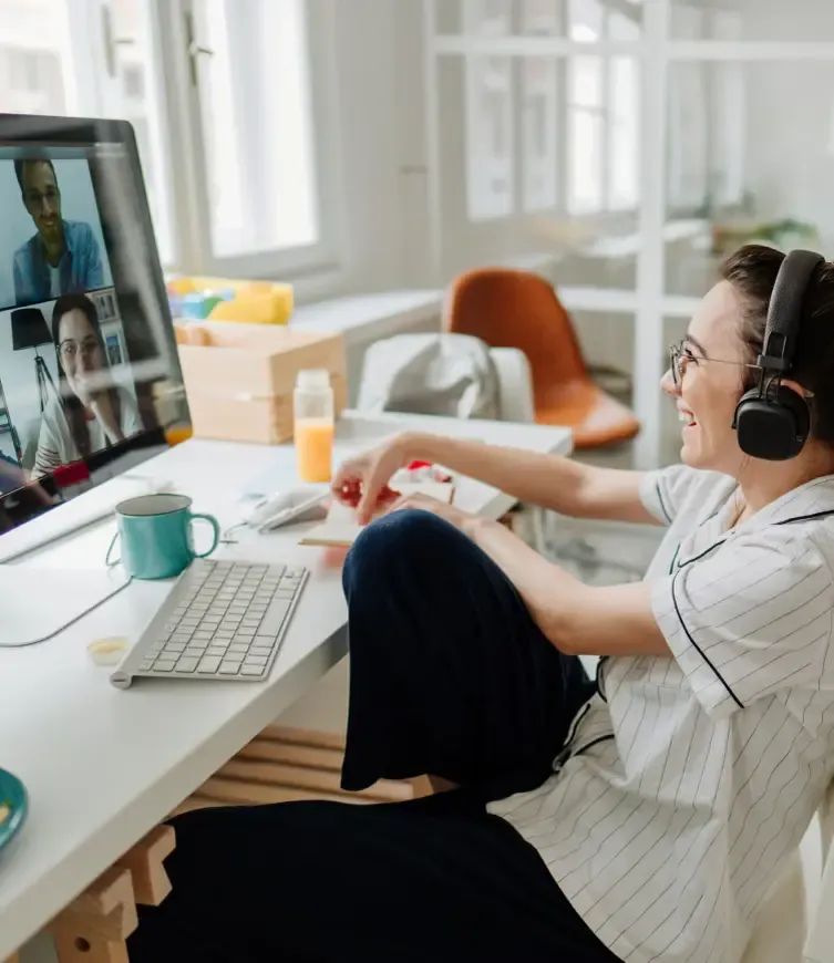 Woman wearing headphones casually sits at desk and laughs while on a virtual call.