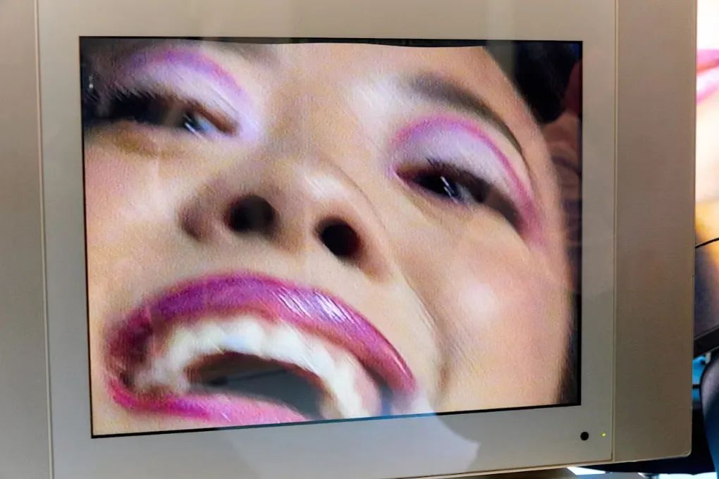 A woman wearing pink eye makeup and pink lipstick is projected on the screen on a mid-1990s model television. She smile's big and is a bit out of focus.
